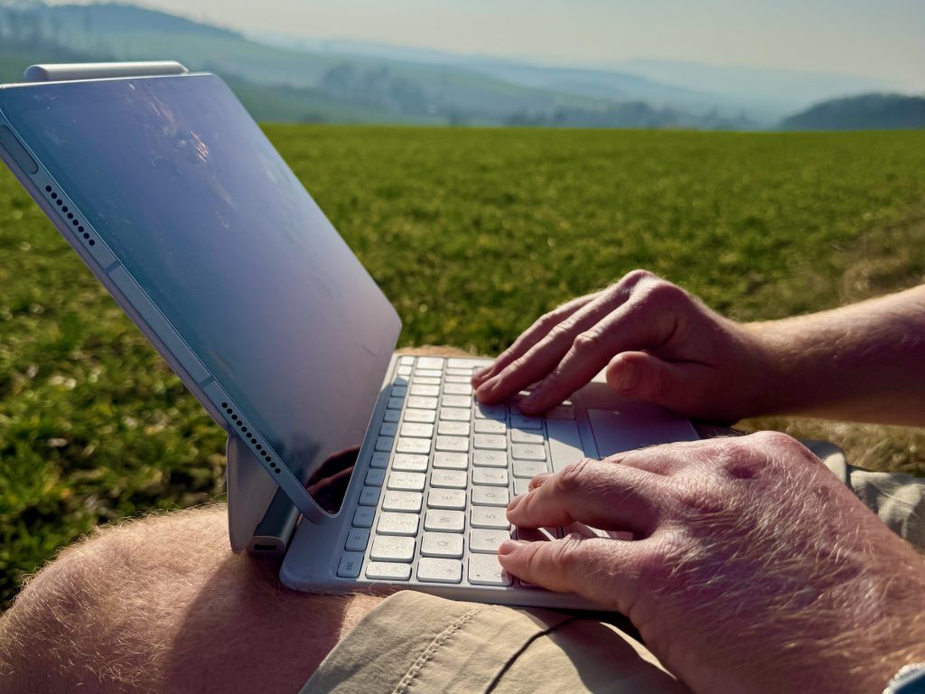 Man sieht auf dem Bild eine Tastatur und Hände, die darauf tippen. Jemand sitzt an einem Feldesrand und tippt auf seine Tastatur. Wenn du an einem ruhigen Ort im Freien arbeiten kannst, hast du die ideale Umgebung für Deep Work.
