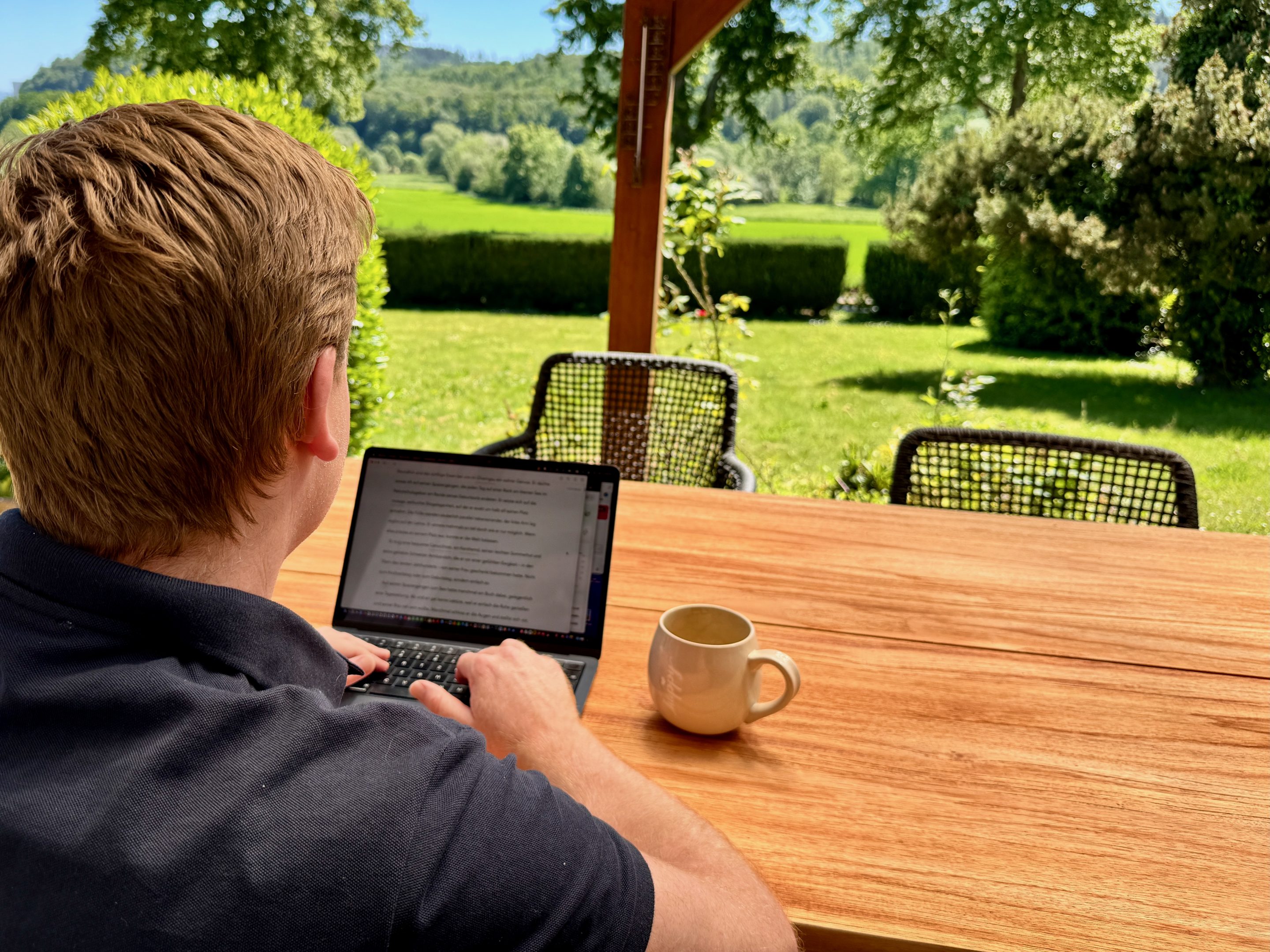 Ein Mann sitzt an einem Tisch und schaut auf einen Computerbildschirm. Neben dem Computer steht eine Tasse mit Kaffee. Der Tisch steht in einem Garten und soll ein Outdoor Office symbolisieren. Die meisten nutzen KI falsch! Lerne mit der "Journalisten-Methode", wie du als Experte mit KI-Strategie überlegene Ergebnisse erzielst.