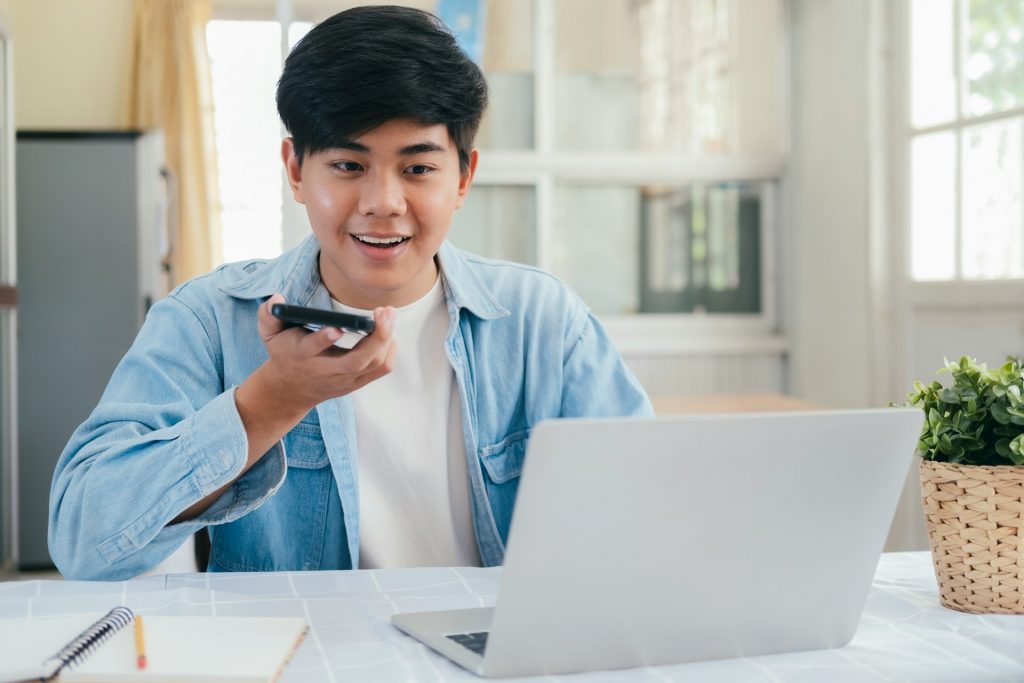Ein Mann diktiert seinen Content-Plan in ein Smartphone und sitzt vor einem Computer. Diktieren beschleunigt die Erstellung deines Content-Plans erheblich.