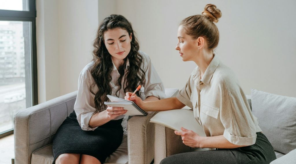 Zwei Frauen sitzen nebeneinander auf Stühlen und arbeiten gemeinsam an etwas. Es sieht so aus, als würden sie einander interviewen. Interviews mit echten Menschen sind Gold wert, um Expertise aus erster Hand zu erhalten – meine "Journalisten-Methode" beschreibt, wie man es richtig angeht.