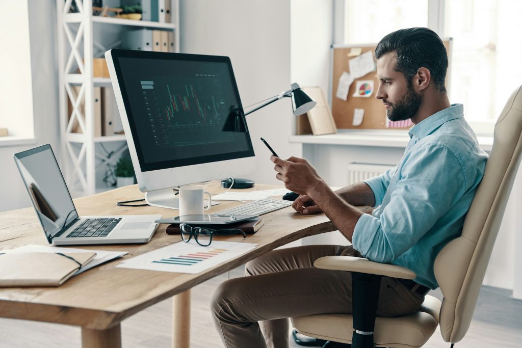 Ein Mann sitzt vor seinem Desktop-Computer und seinem Laptop am Schreibtisch und schaut auf sein Smartphone.
Wenn du kognitive Verzerrungen bei dir erkennst, kannst du bewusst gegensteuern und eine objektivere Entscheidung treffen.