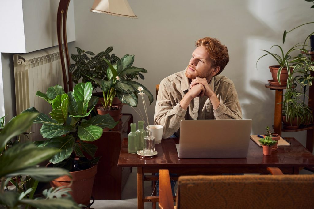 Ein Mann sitzt an einem Schreibtisch vor seinem Laptop und blickt in die Luft. Er scheint intensiv nachzudenken. Auf der anderen Seite kann reines Remote-Arbeiten dich in die Isolation treiben. Wenn du sechs Stunden am Tag in Deep Work versinkst, hast du keine Zeit und auch nicht das Bedürfnis für soziale Interaktion.