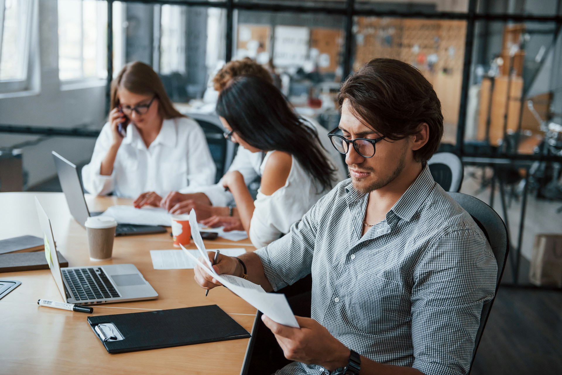 Auf dem Bild sieht man einen Mann, der weiter vorne vor einem Computer sitzt und im Hintergrund sitzen drei seiner Arbeitskolleginnen vor einem Computer. Um den Mann vorne könnte es sich um einen stillen Experten handeln, der für sich alleine arbeitet, während die anderen im Team arbeiten.
