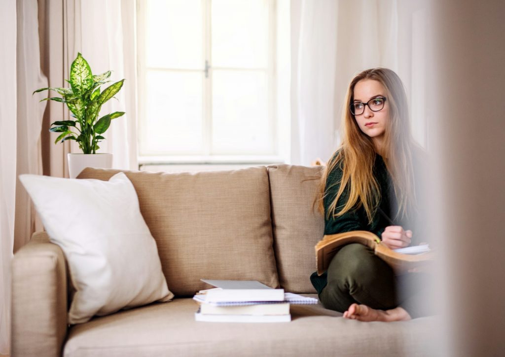 Eine Frau mit Brille sitzt auf einem Sofa und denkt augenscheinlich nach, während sie von mehreren Büchern umgeben ist. Bist du eine stille Expertin? Das hat seine Vor- und Nachteile. Ich zeige dir, wie du diese Gabe zu deinem Vorteil nutzt.