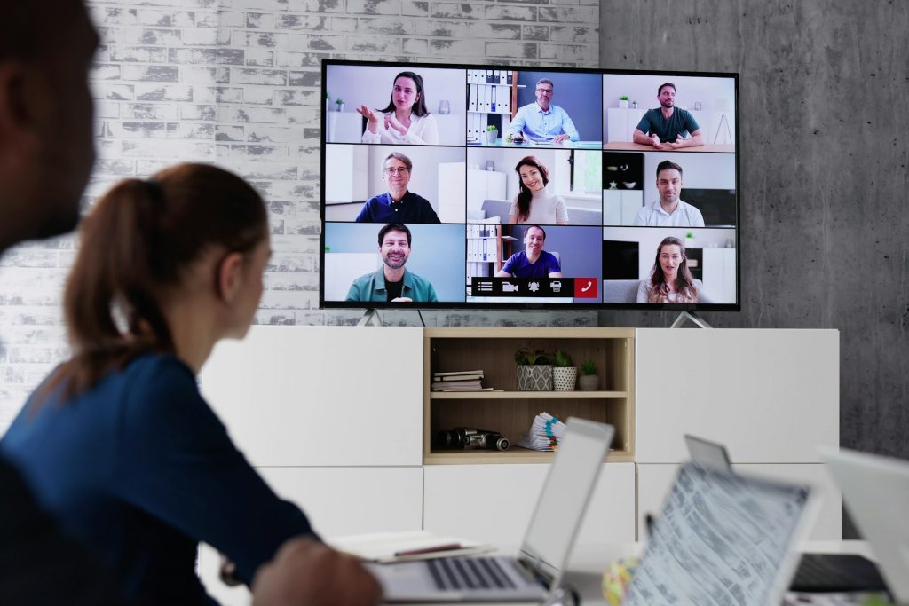 Auf dem Bild sieht man einen Meetingraum. An einem Tisch sitzen Leute vor ihren Computern und auf einem großen Fernseher sind andere Leute in kleinen Kacheln aufgezeigt. Es findet also eine große Videokonferenz statt mit vielen Teilnehmern.
Schmerz lasst nach! Kaum jemand ist ein ernsthafter Fan von Videotelefonaten – insbesondere in Gruppen.
